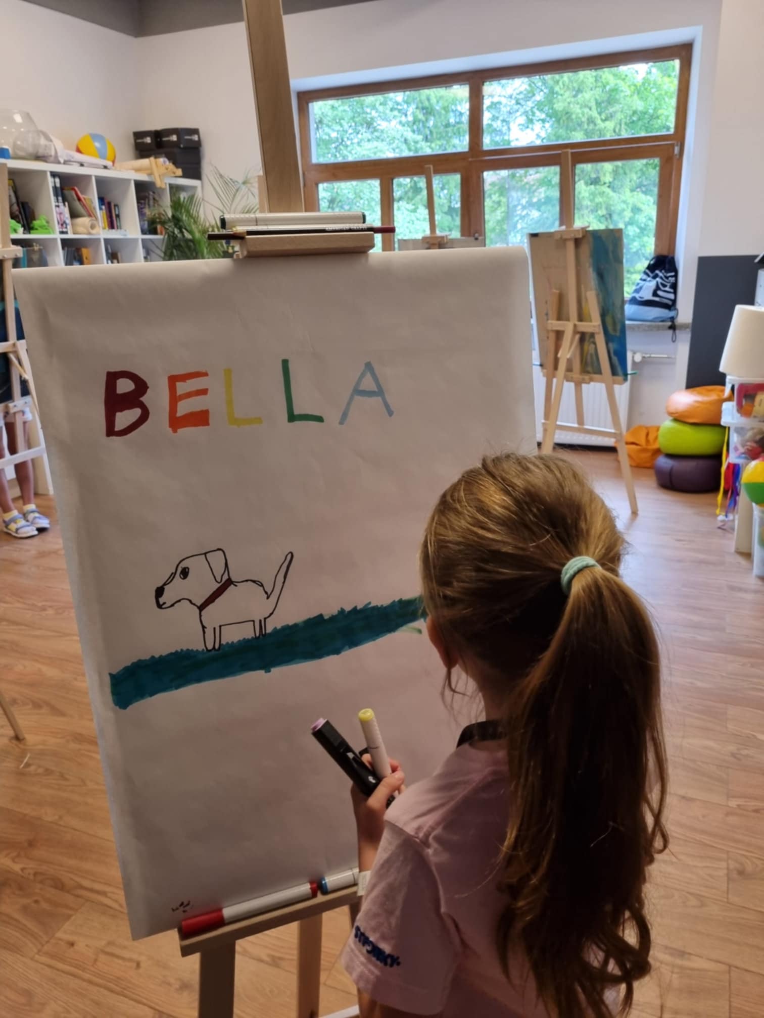 Dziewczynka stoi przed sztalugą, na której jest rysunek psa i napis Bella