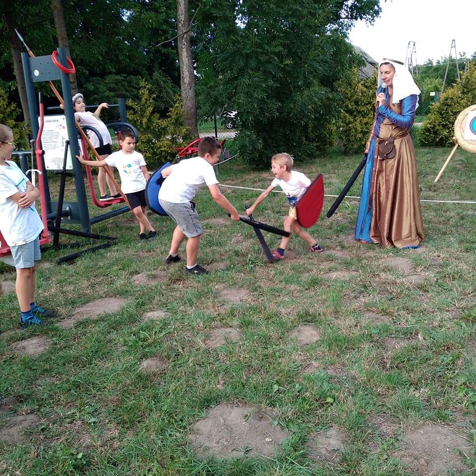 Dwóch chłopców walczących w rycerskim turnieju. Obok nich stoi kobieta w przebraniu, przypominającym średniowieczny strój.