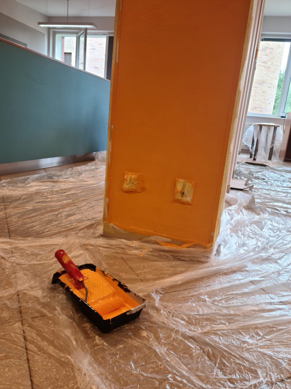 Widok na remontowaną salę - w tle niskie niebieskie ścianki tzw. boxów. Na pierwszym planie fragment ściany pomalowanej na pomarańczowo, na podłodze rozłożona folia malarska i narzędzia malarskie.