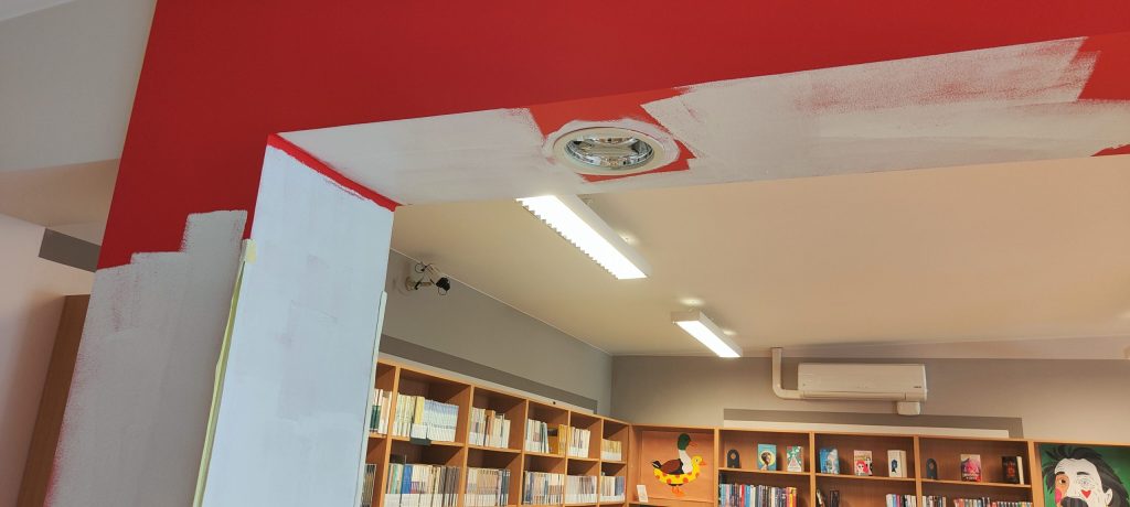 Widok na fragment malowanej ściany z koloru czerwonego na biały. W tle regały z książki.