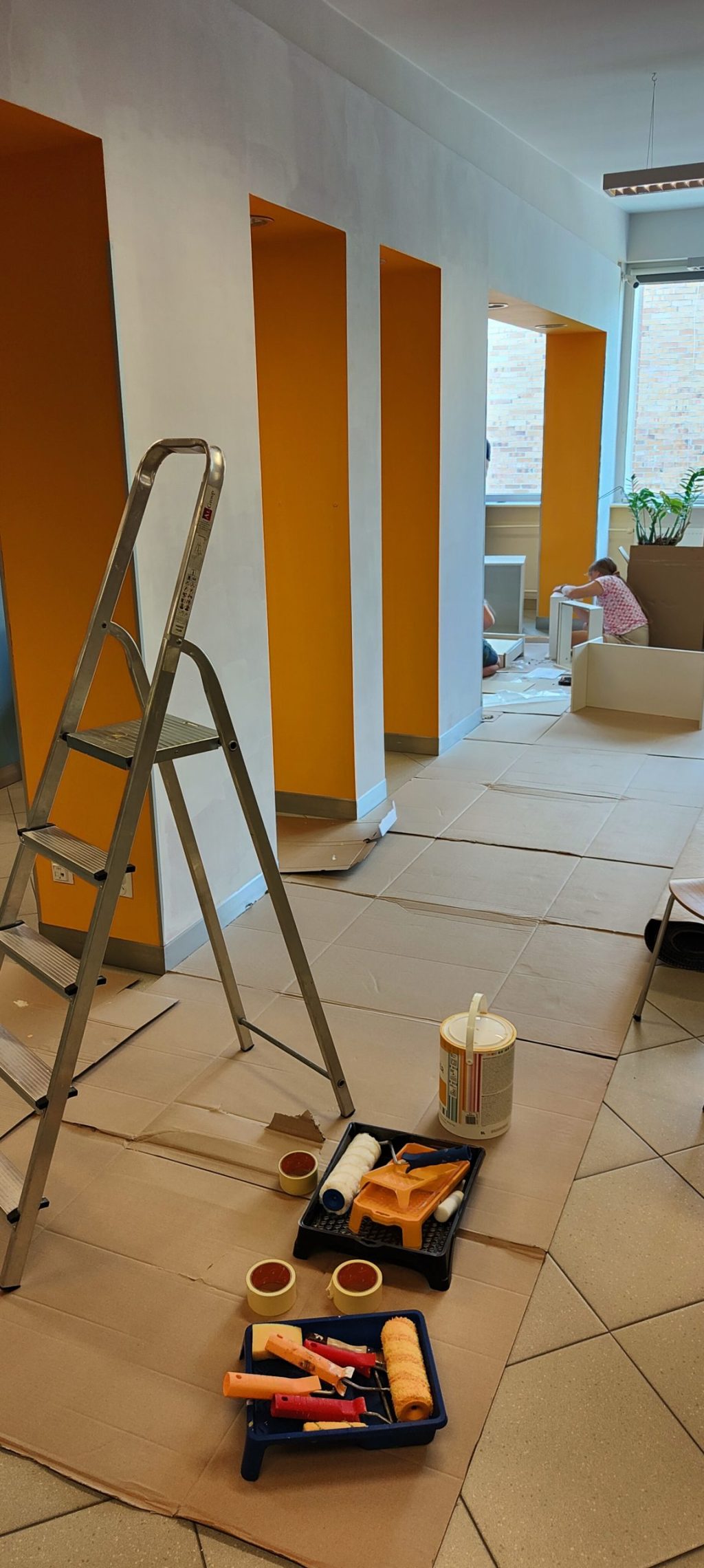 Widok na remontowane pomieszczenie - po prawej stronie łuk ścienny pomalowany na pomarańczowo i biało, w tle kucająca dziewczyna. Na pierwszym planie drabina i farba. Na podłodze karton.