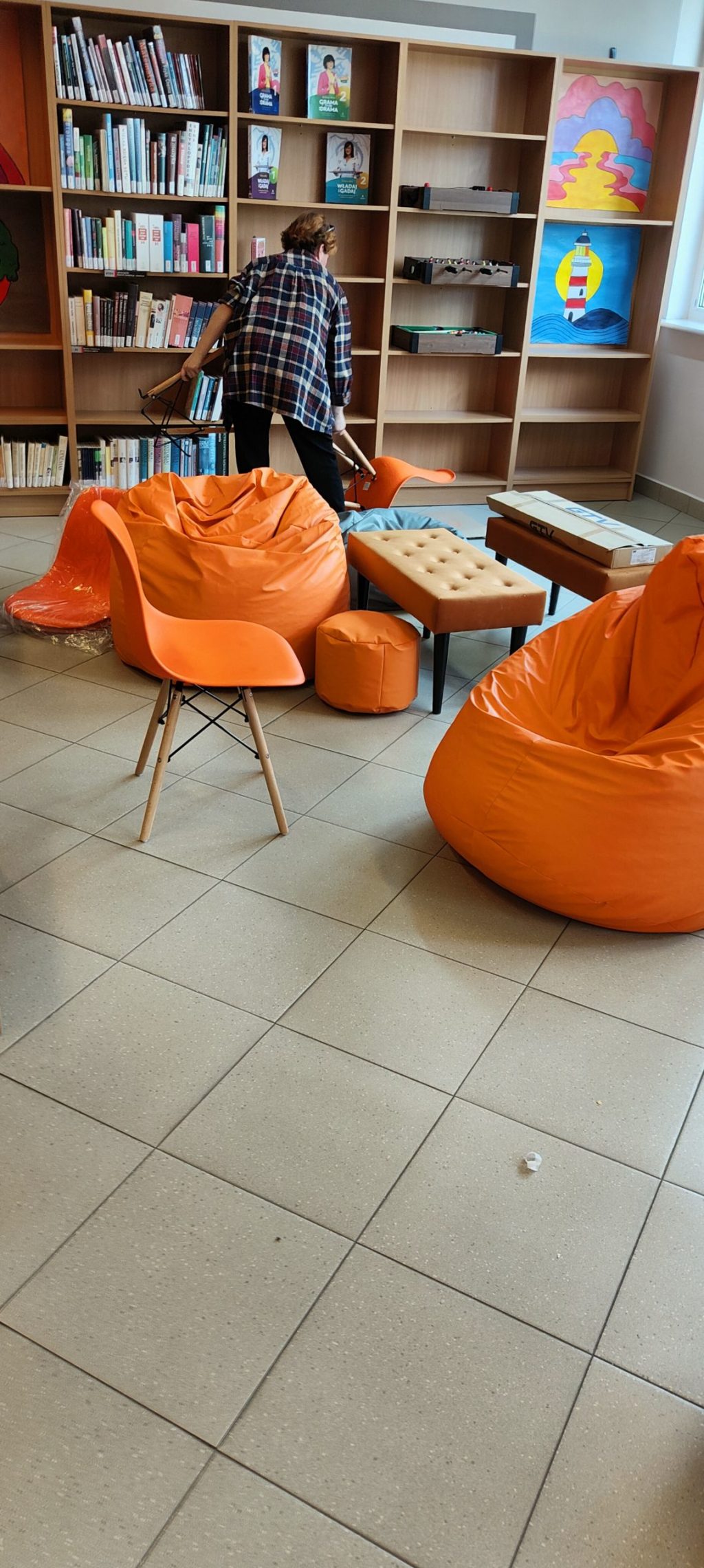 Zdjęcie przedstawiające elementy umeblowania - pomarańczowe krzesło i poduchy, ławki tapicerowane. W tle regały z książkami i niosąca krzesło kobieta.
