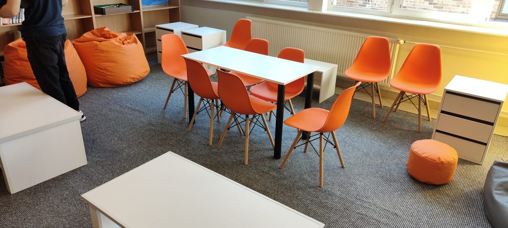 Zdjęcie mebli - biały stół i pomarańczowe krzesła, w tle białe kontenerki i stolik.