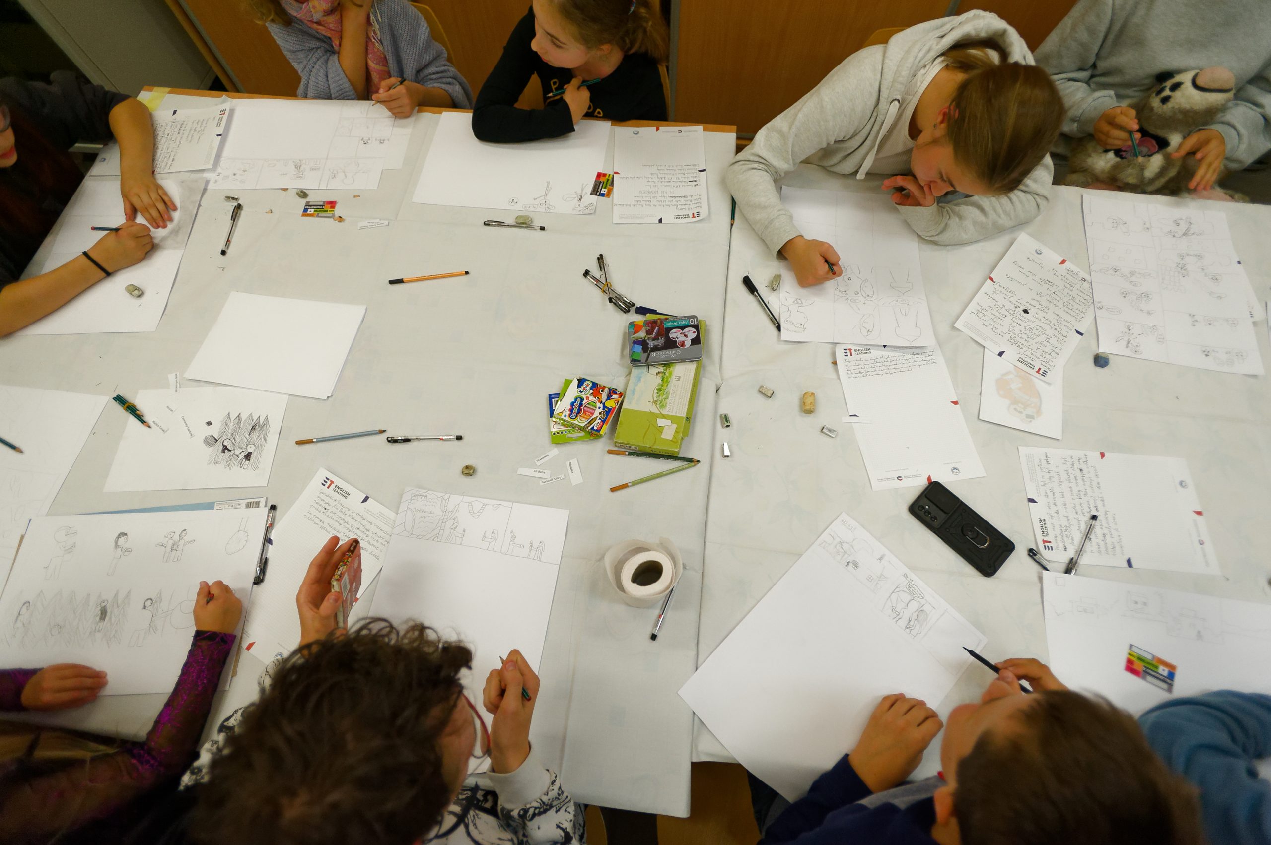 Widok z góry na grupę dzieci siedzących przy stole i rysujących na białych kartkach. Między kartkami leżą przybory plastyczne.