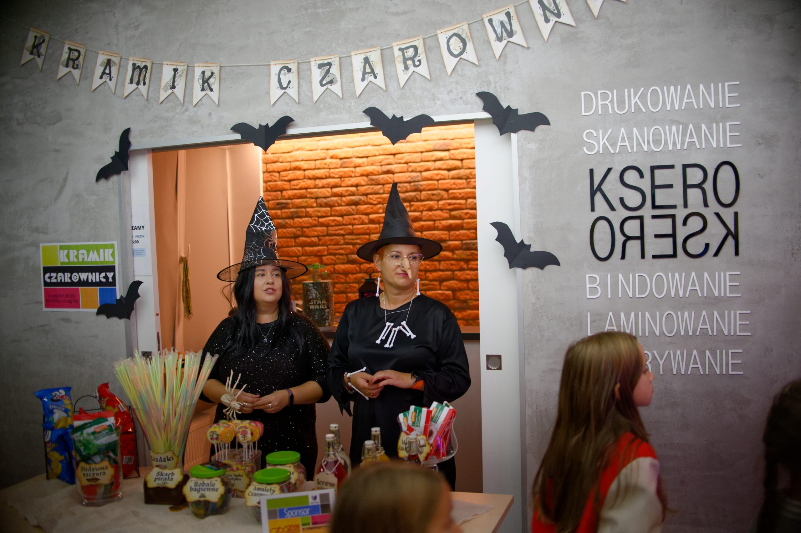 Dwie kobiety przebrane za czarownice stojące w wejściu do punktu ksero. Przed nimi stoi stół zastawiony słoikami ze słodyczami i butelkami z napojami.
