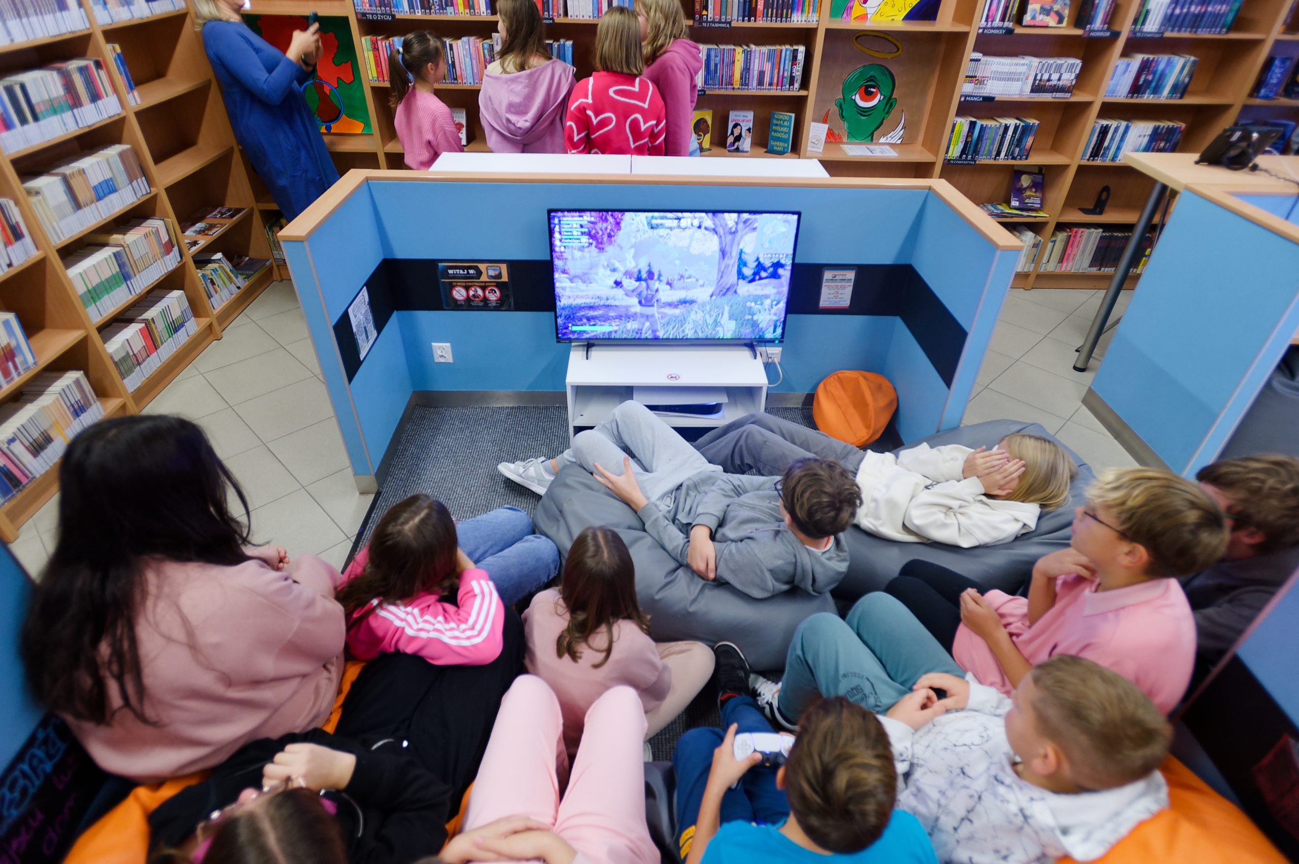 Grupa nastolatków siedząca przed telewizorem, w tle osoby stojące przy regałach z książkami.