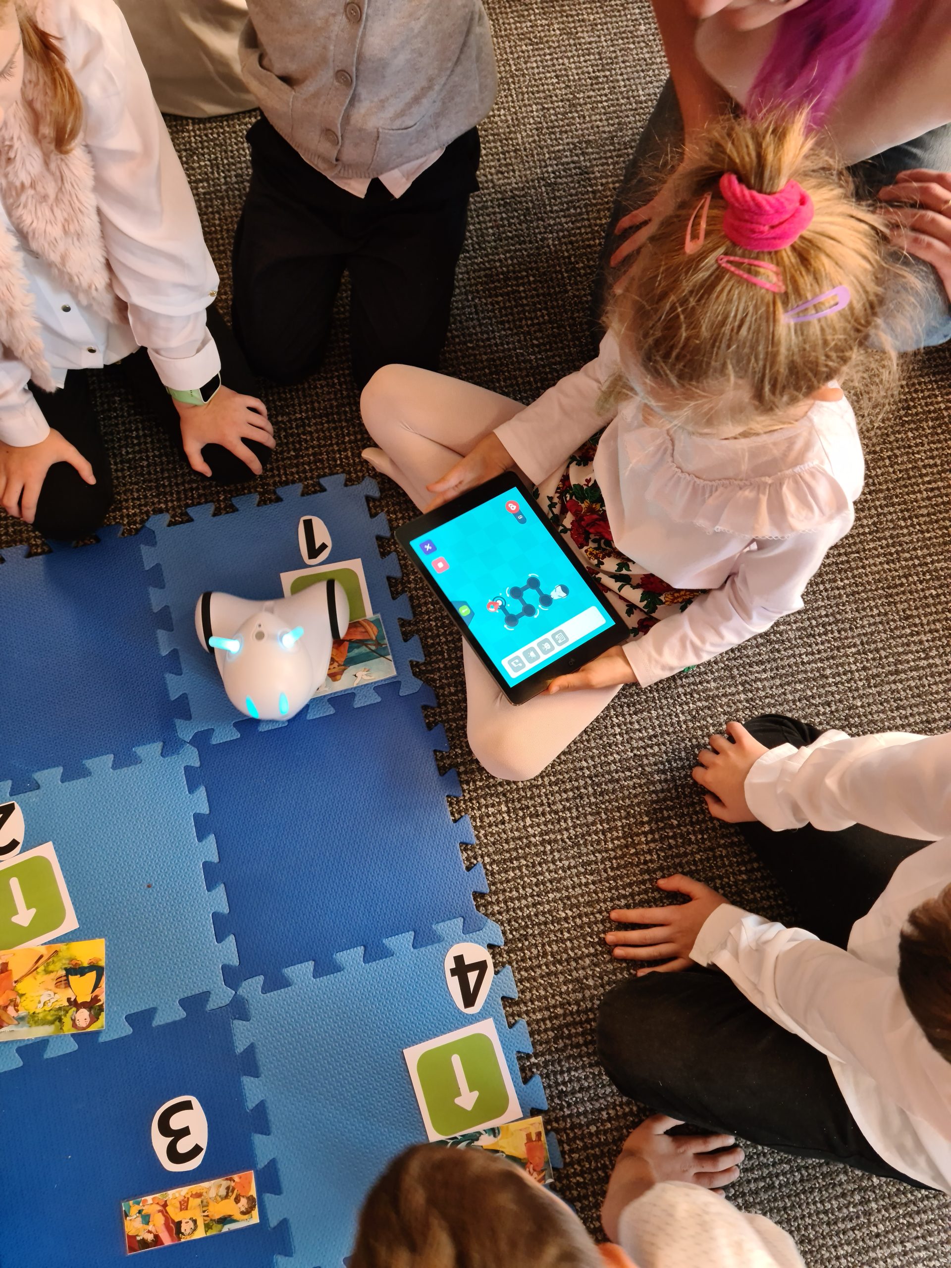 Dzieci siedzące wokół niebieskiej maty na której stoi robot. Jedno z dzieci trzyma w ręku tableta.