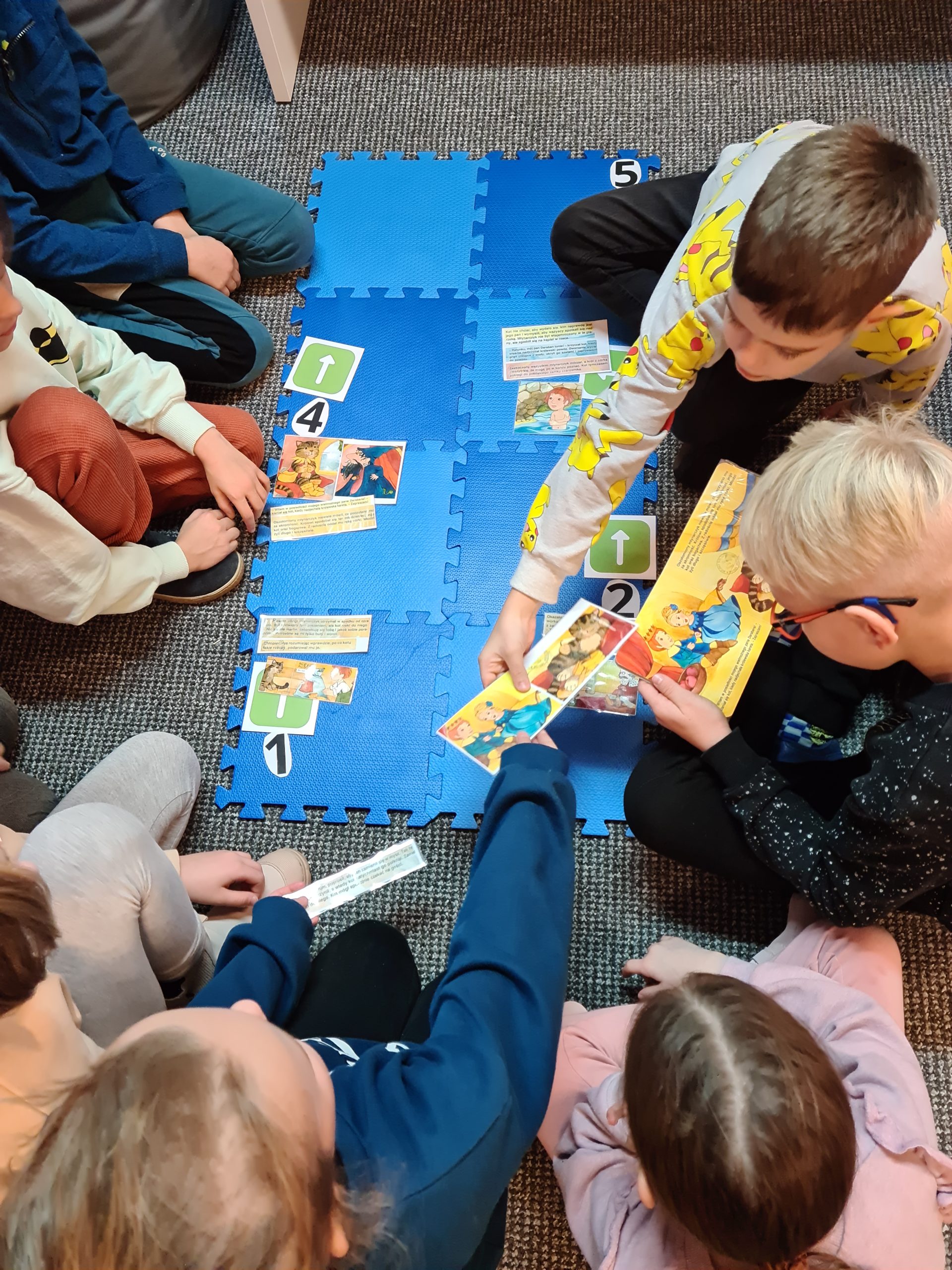 Dzieci siedzące w grupie na podłodze. Jedno dziecko trzyma otwartą książkę, inny chłopiec trzyma w ręku ilustrację.