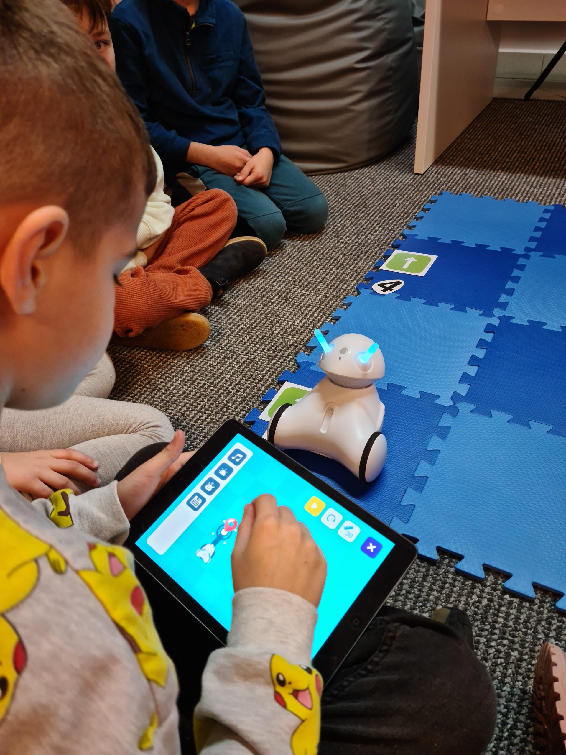 Dzieci siedzące wokół niebieskiej maty na której stoi robot. Jedno z dzieci trzyma w ręku tableta.