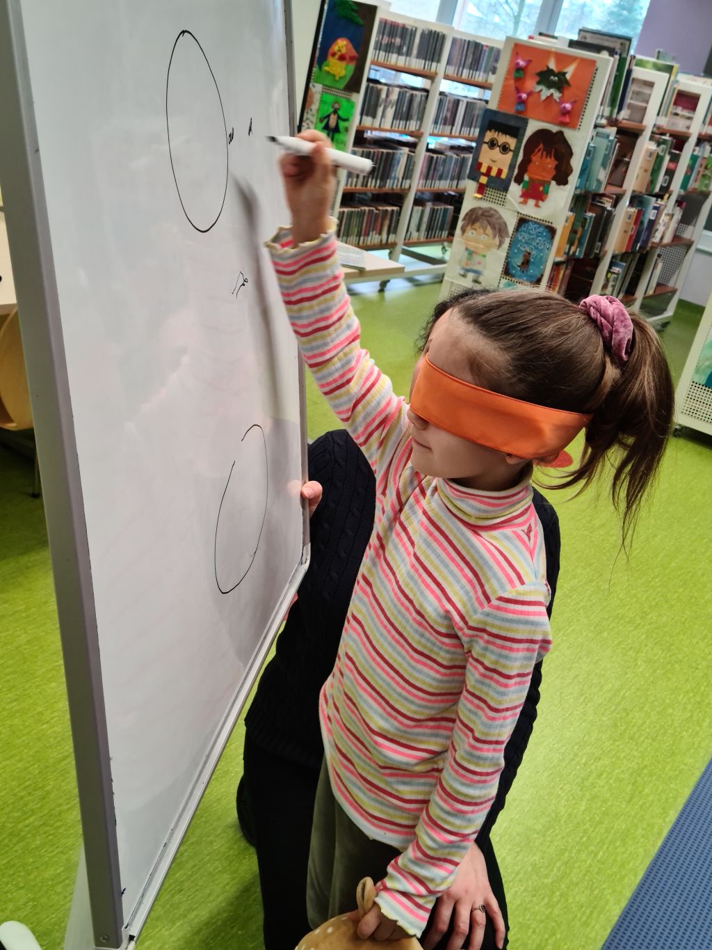 Dziecko z opaską na oczach rysuje markerem po tablicy.