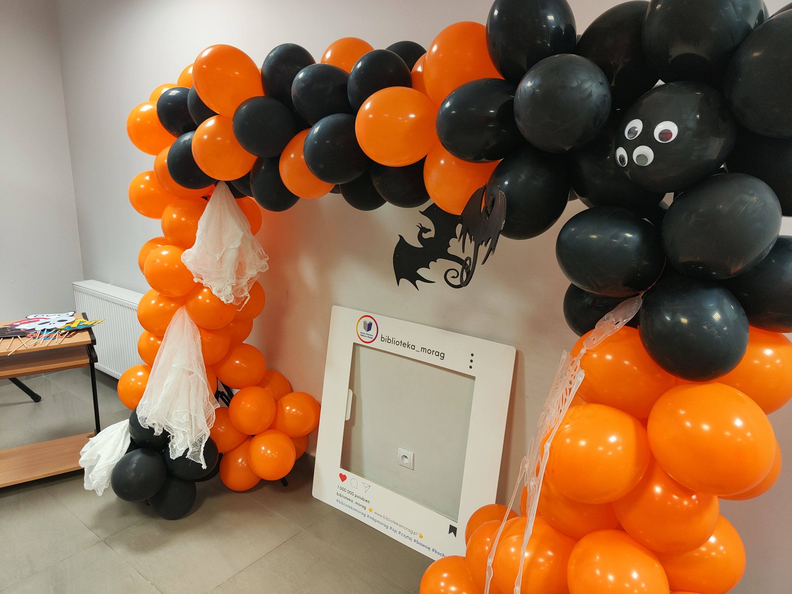 Łuk z czarnych i pomarańczowych balonów, na środku tekturowa ramka do zdjęć.