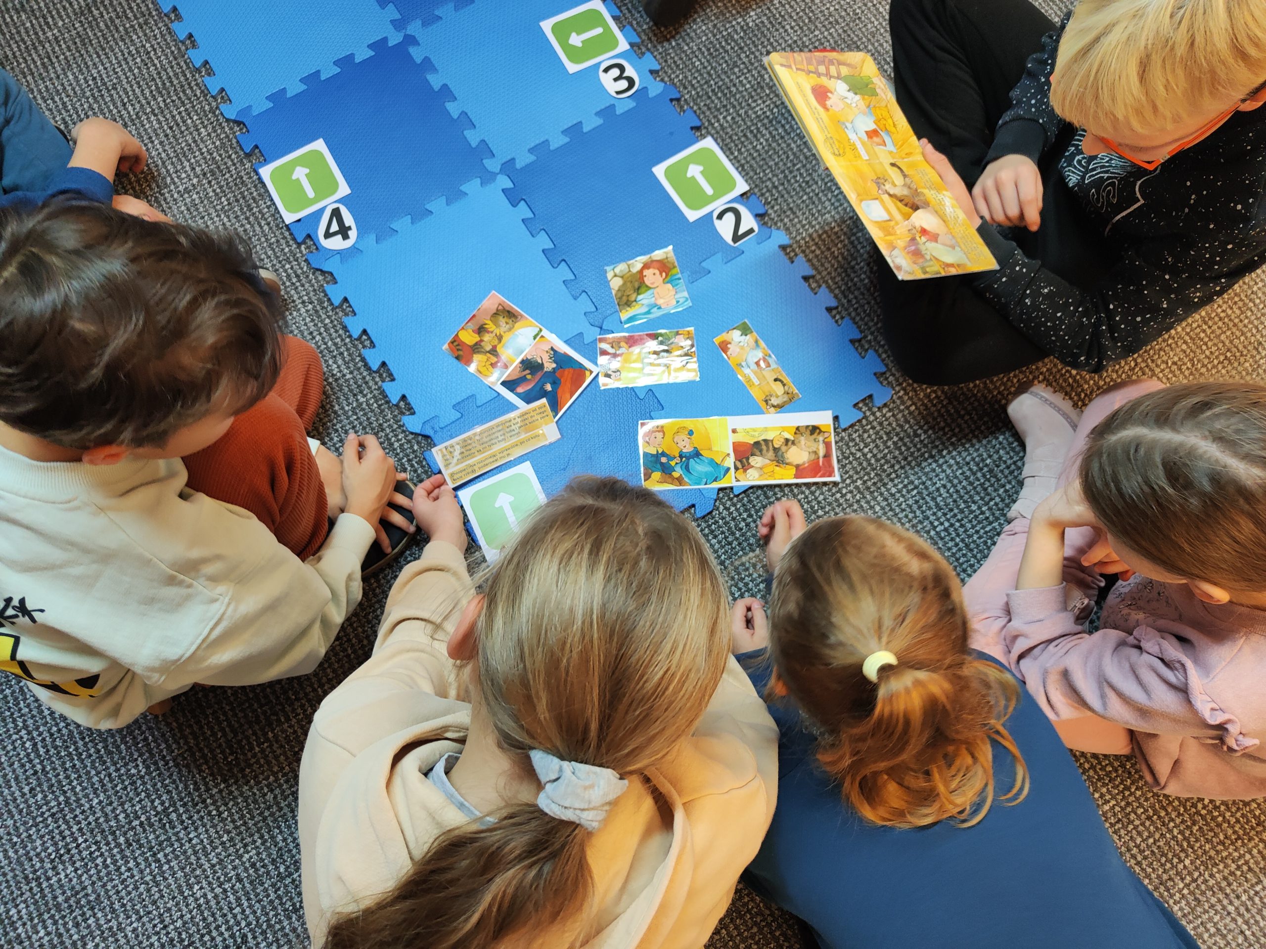 Dzieci siedzące w grupie na podłodze pochylające się nad książką i ilustracjami.