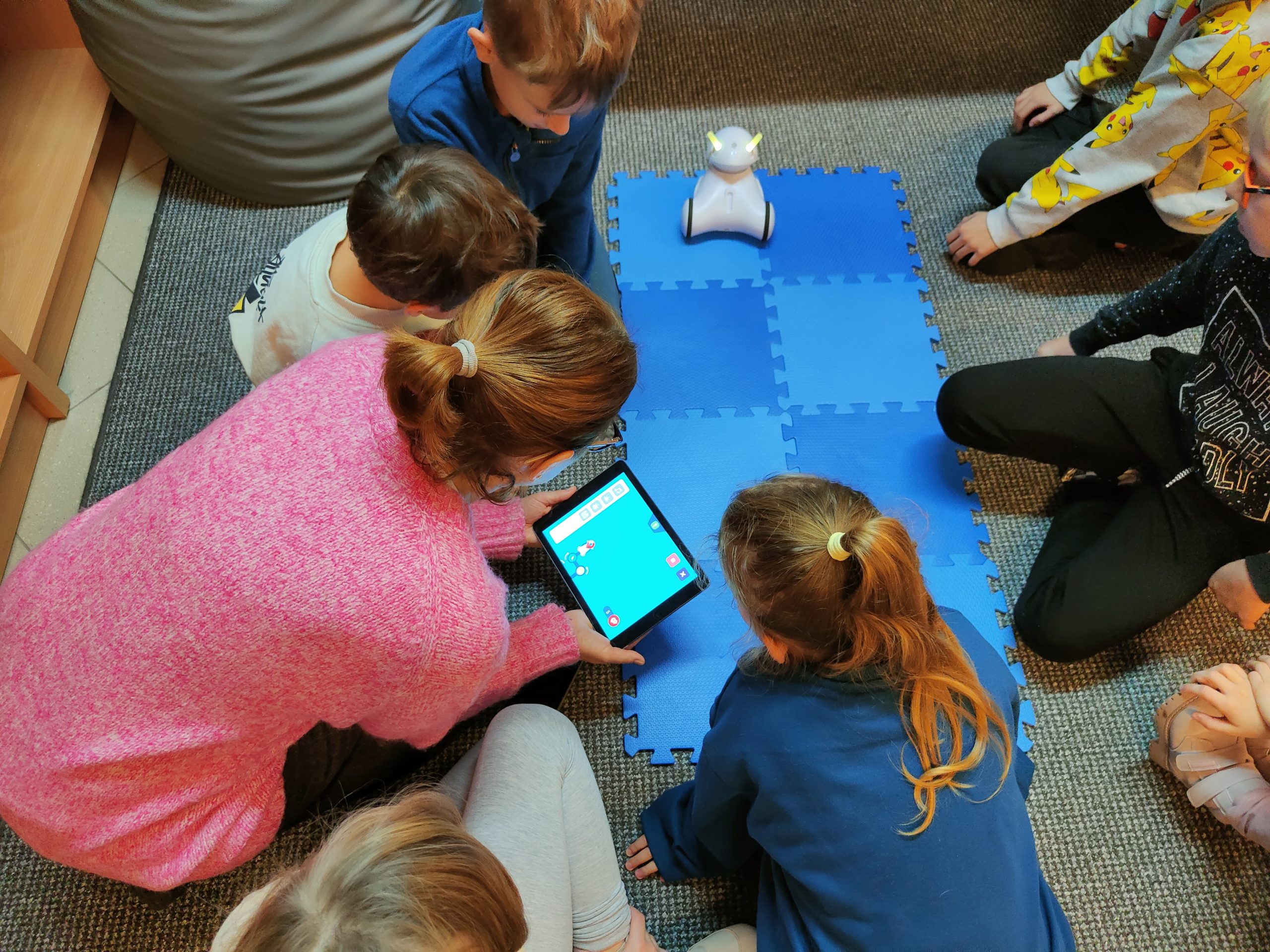 Dzieci siedzące na podłodze z dorosłą kobietą trzymającą tablet.
