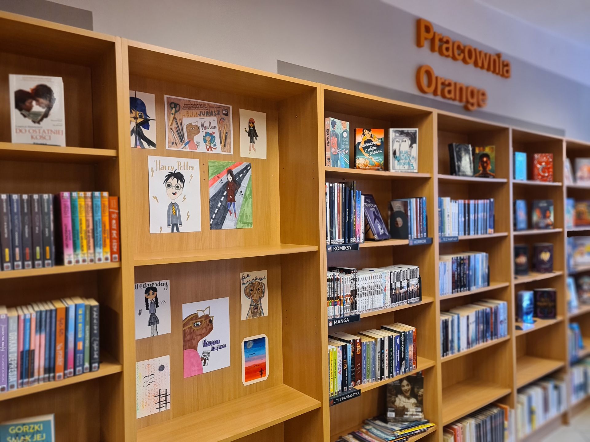 regały z książkami pod ścianą, z jednego z nich są wyjęte półki, a w wolnym miejscu wiszą prace plastyczne, nad regałami logo pracowni Orange