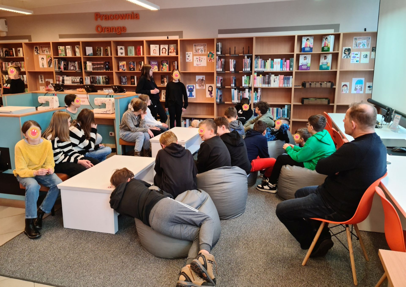 Grupa uczniów siedzi na pufach i wraz z nauczycielem zadają pytania młodej artystce