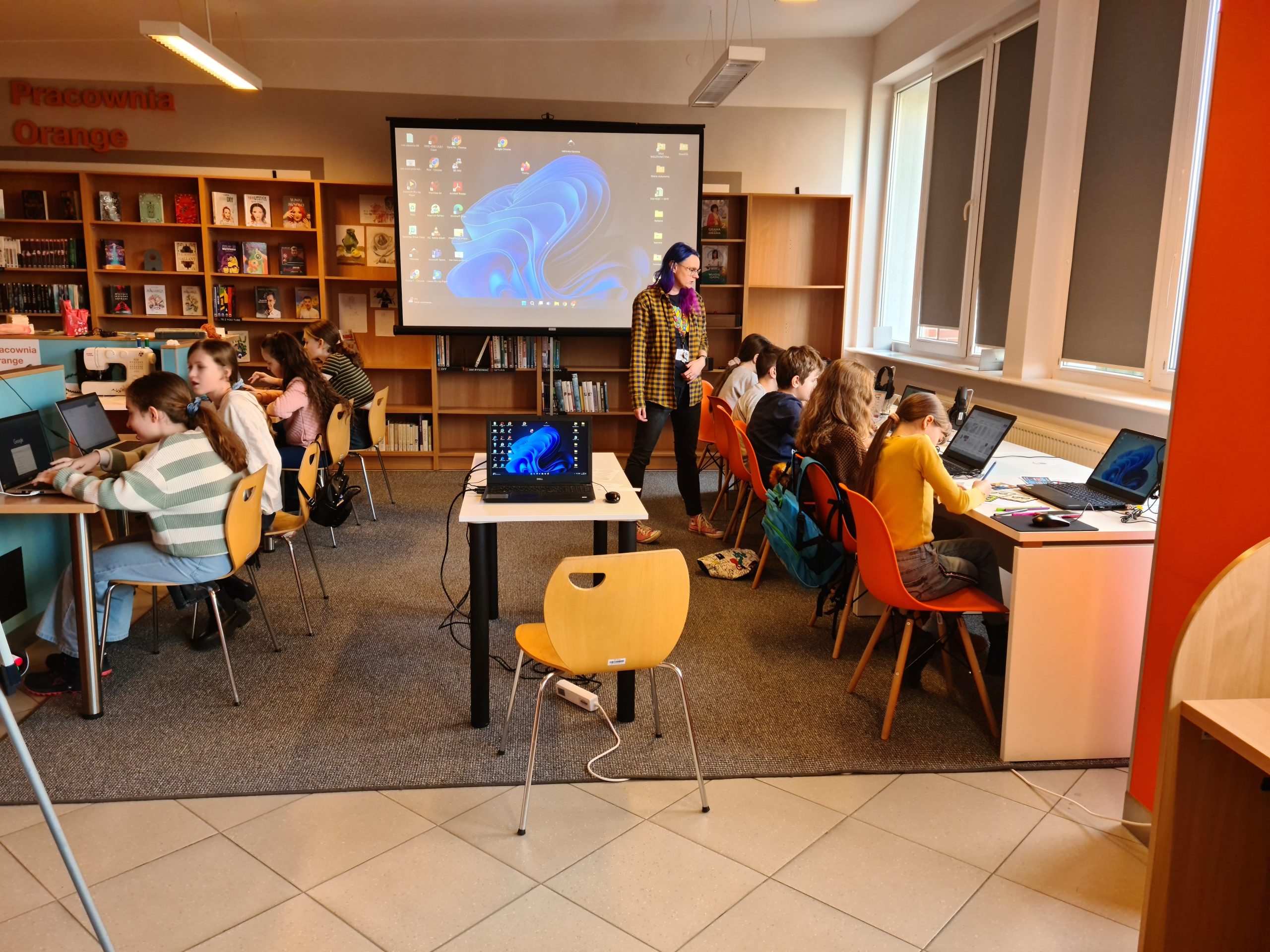 Grupa dzieci siedząca przy biurkach z laptopami, po środku stoi dorosła kobieta i biurko z laptopem, w tle ekran projekcyjny.