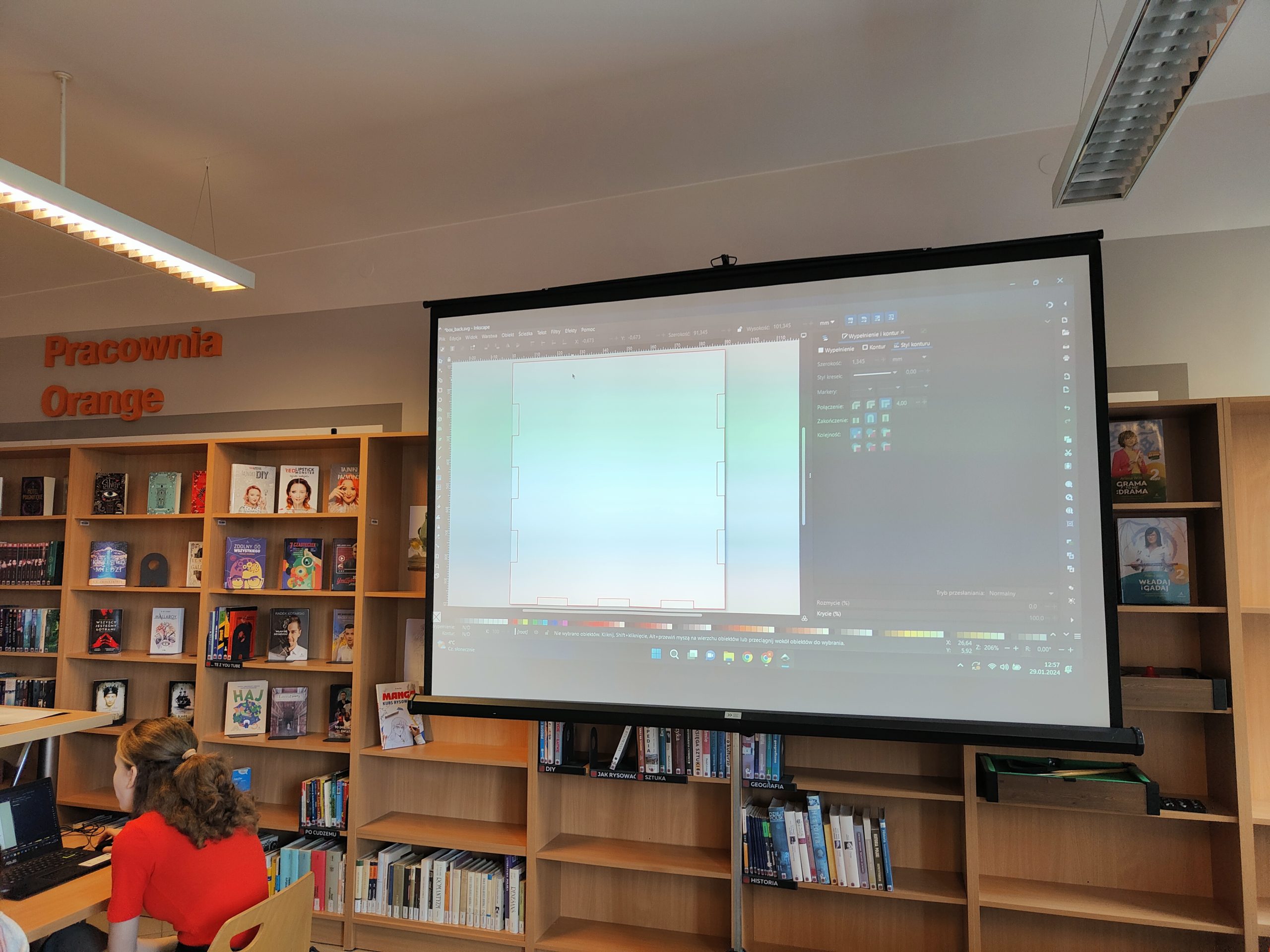 Ekran z wyświetlanym obrazem z projektora, stojący na tle regałów z książkami.
