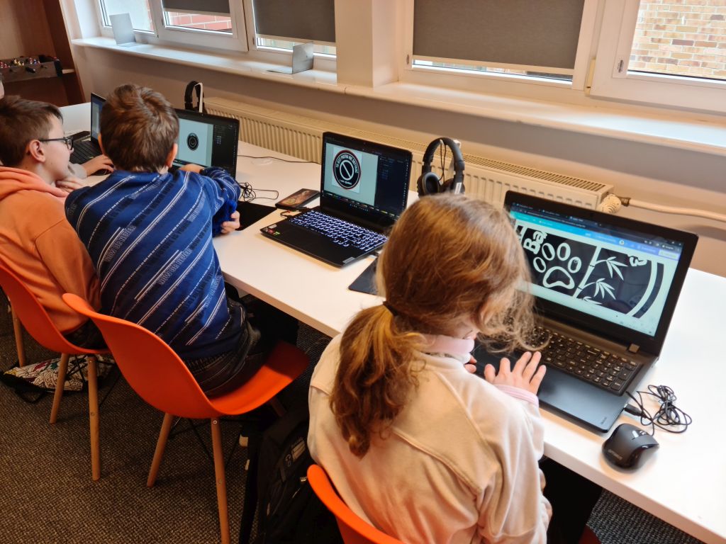 Młodzież siedzi przy biurkach i korzysta z laptopów.