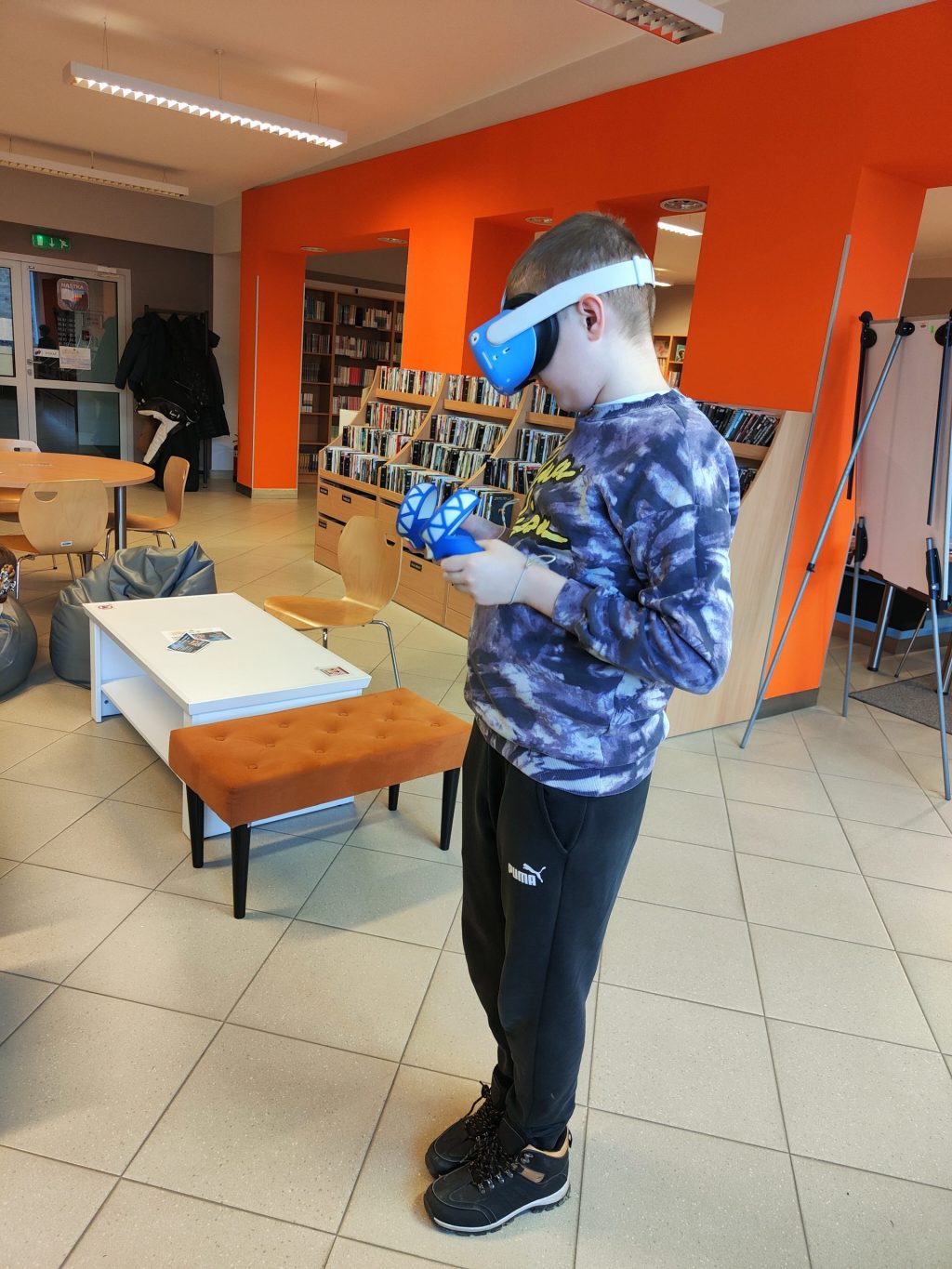 Chłopiec korzystający z gogli VR. W tle regały z filmami, stolik i pufa.