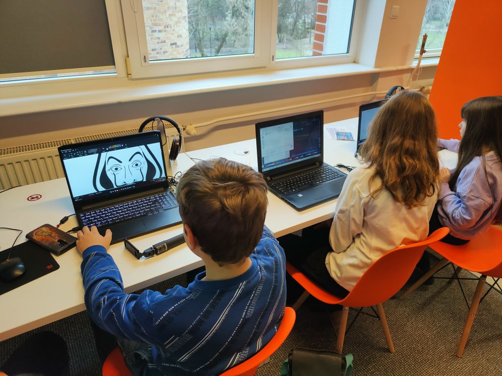Młodzież siedzi przy biurkach i korzysta z laptopów.