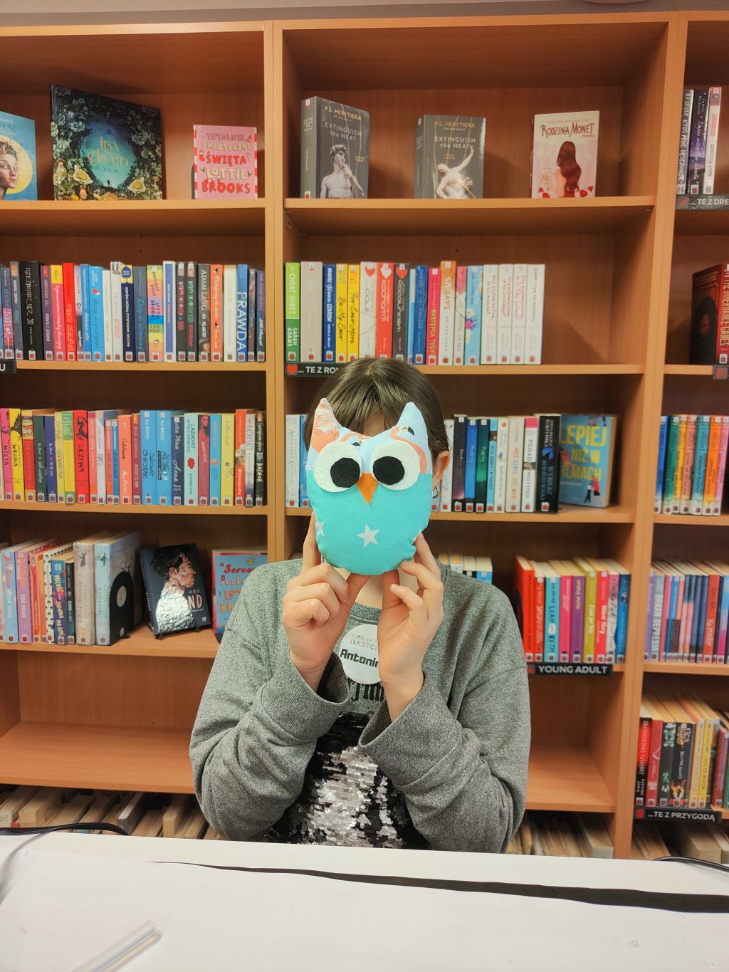 Dziewczynka stoi na tle regałów z książkami trzymając przed twarzą pluszową niebieską sowę.
