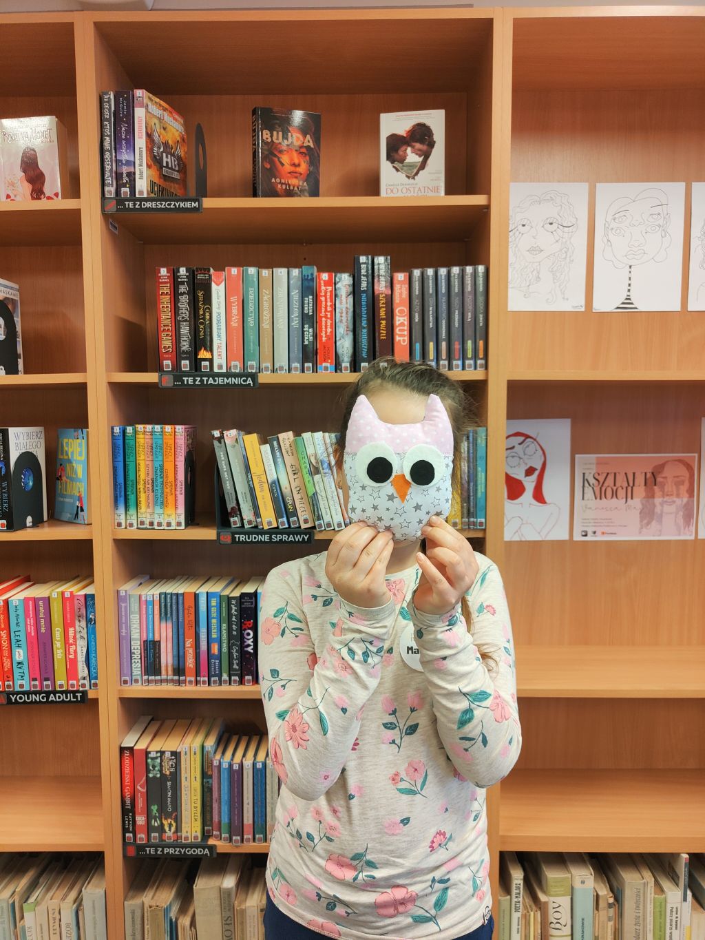 Dziewczynka stoi na tle regałów z książkami trzymając przed twarzą pluszową siwo-różową sowę.