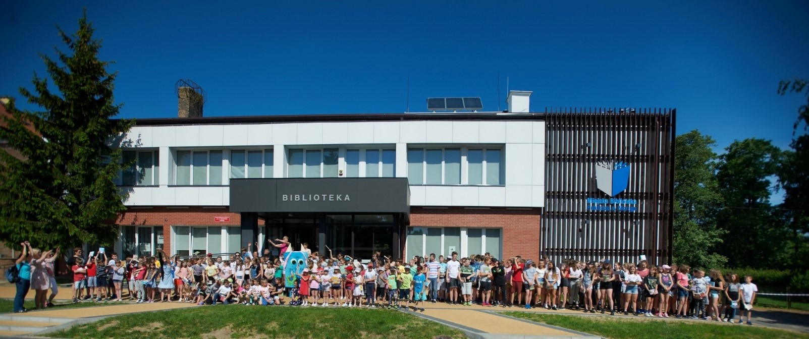 Duża grupa dzieci i maskotka Czytosław stoją przed budynkiem biblioteki