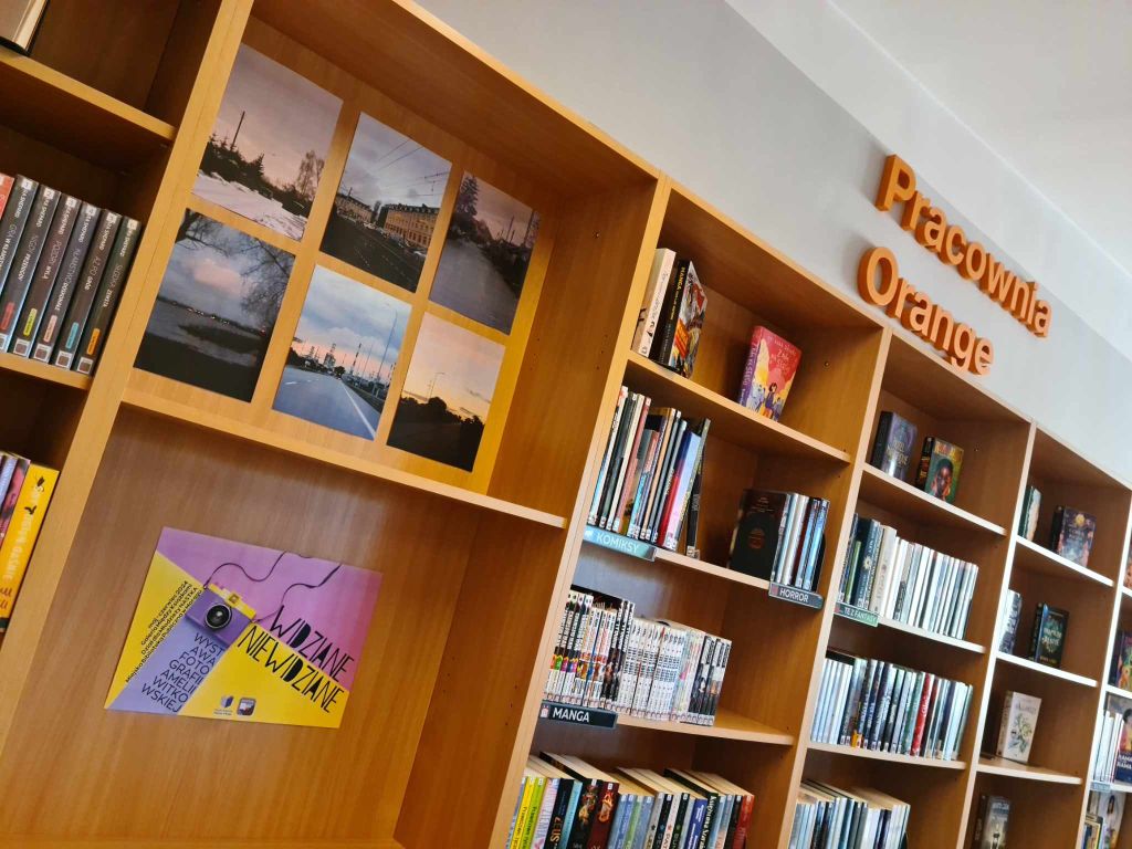 Regał z książkami po prawej, po lewej na tylną ścianę regału wklejone zdjęcia i plakat, nad regałami pomarańczowy napis Pracownia Orange