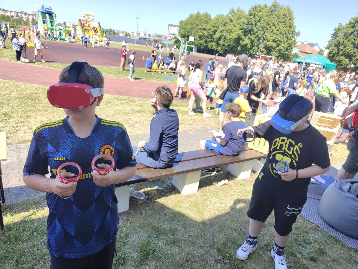 dwóch chłopców korzysta z gogli wirtualnej rzeczywistości na dworze, w tle boisko szkolne i dzieci biorące udział w festynie
