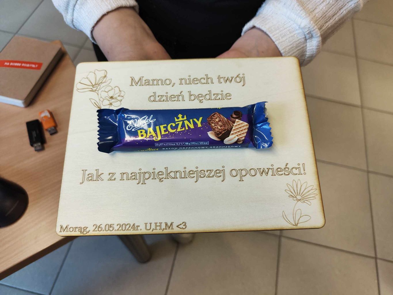 Tabliczka wycięta ze sklejki z życzeniami na Dzień Matki, ozdobną grafiką i batonem Bajeczny przyklejonym na środku.