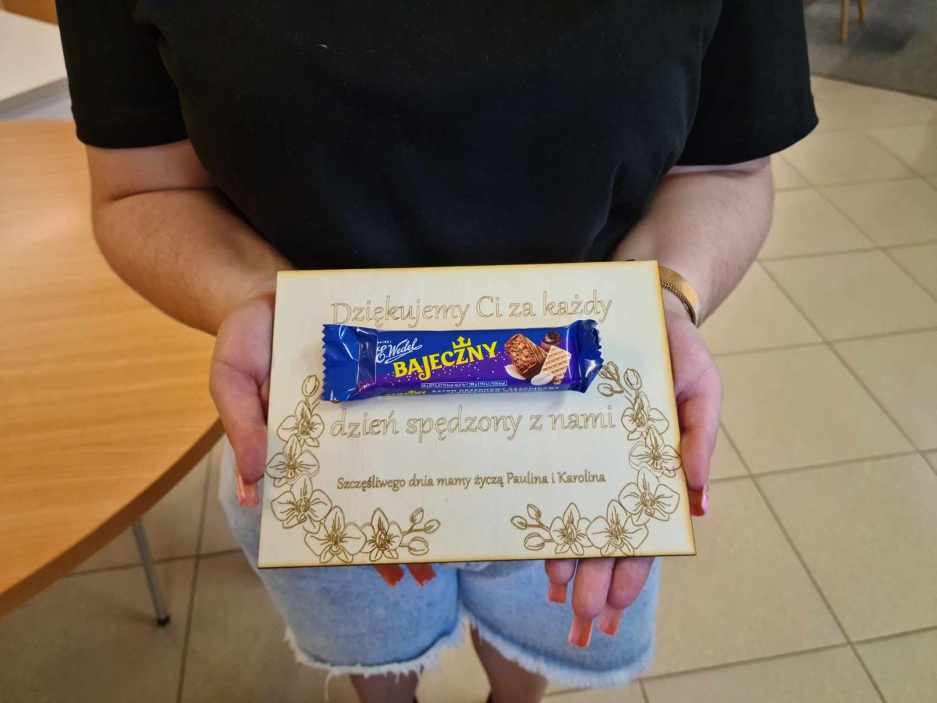 Tabliczka wycięta ze sklejki z życzeniami na Dzień Matki, ozdobną grafiką i batonem Bajeczny przyklejonym na środku.
