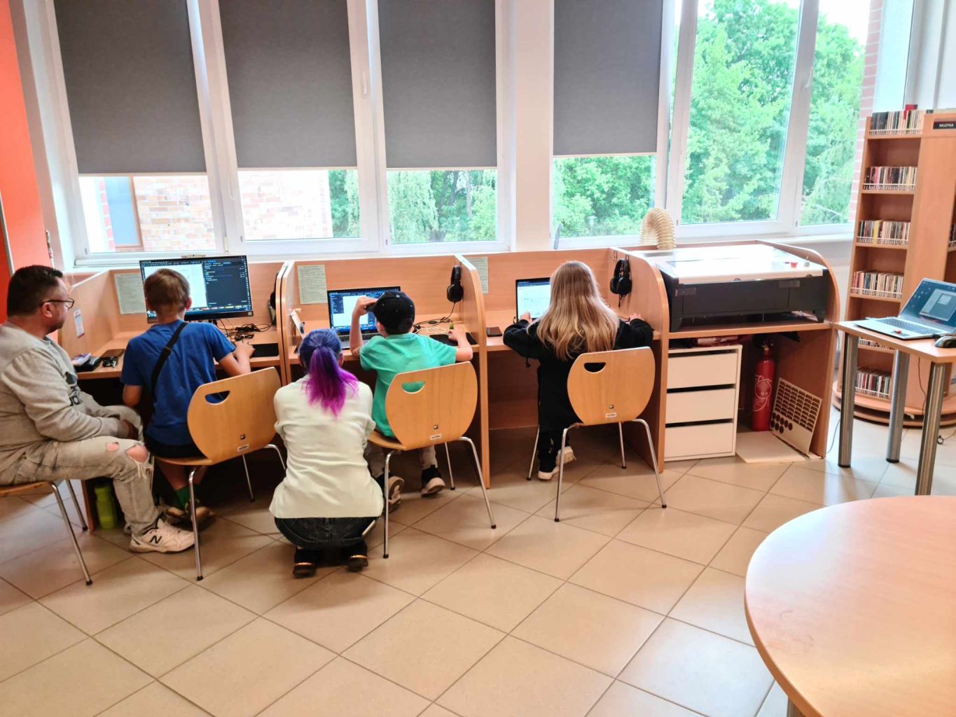 Grupa dzieci siedzi przy komputerach. Obok jednego z chłopców kuca kobieta.