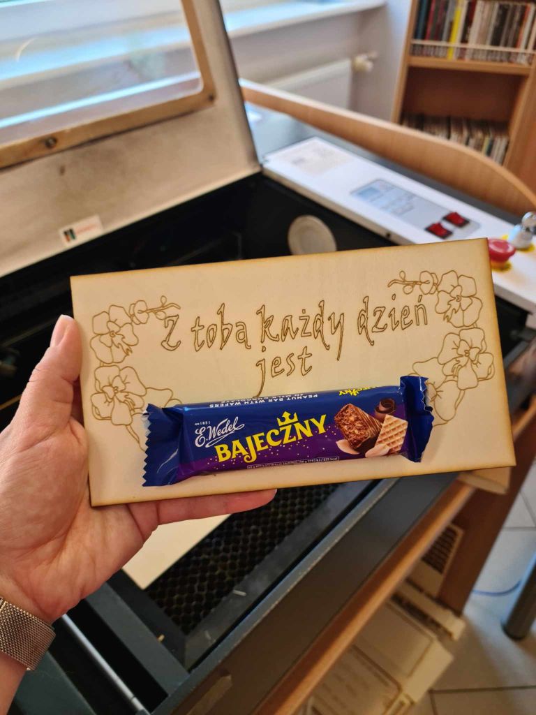 Tabliczka wycięta ze sklejki z życzeniami na Dzień Matki, ozdobną grafiką i batonem Bajeczny przyklejonym na środku.