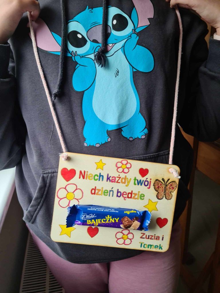 Tabliczka wycięta ze sklejki z życzeniami na Dzień Matki, ozdobną grafiką i batonem Bajeczny przyklejonym na środku.