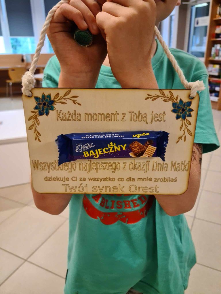 Tabliczka wycięta ze sklejki z życzeniami na Dzień Matki, ozdobną grafiką i batonem Bajeczny przyklejonym na środku.