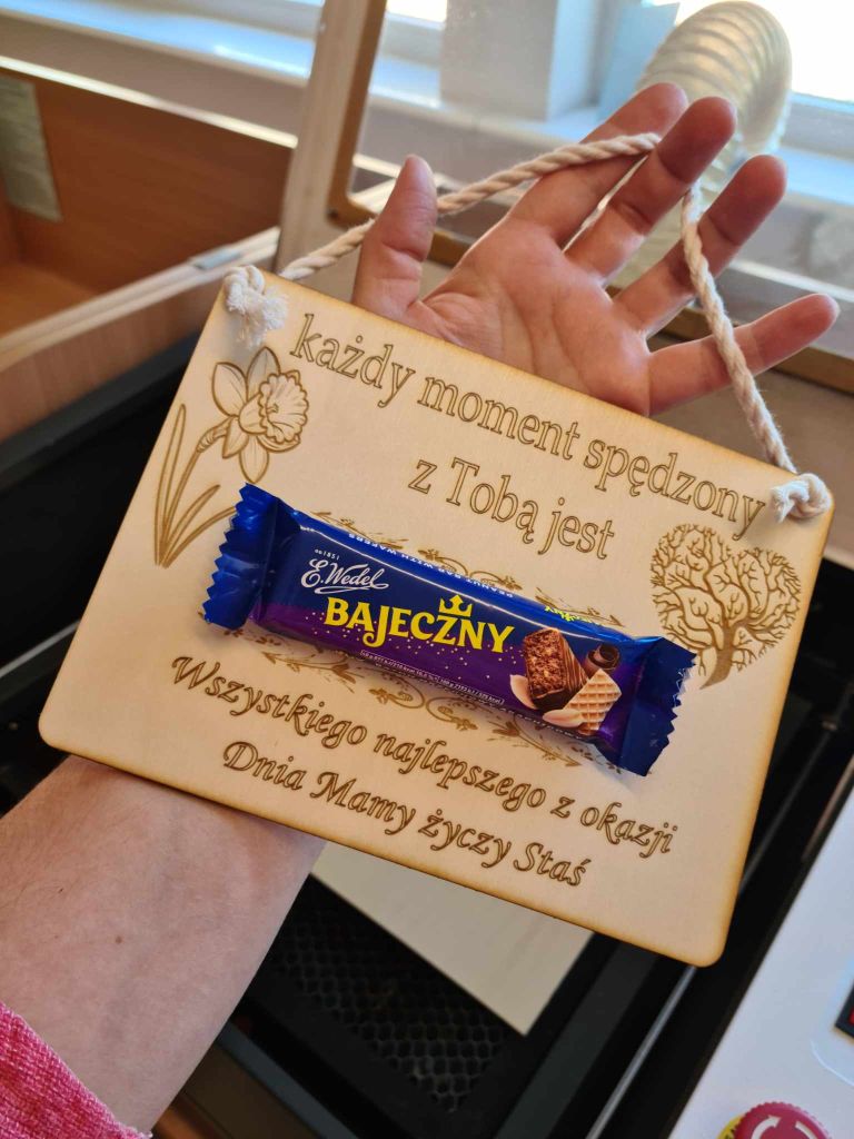 Tabliczka wycięta ze sklejki z życzeniami na Dzień Matki, ozdobną grafiką i batonem Bajeczny przyklejonym na środku.