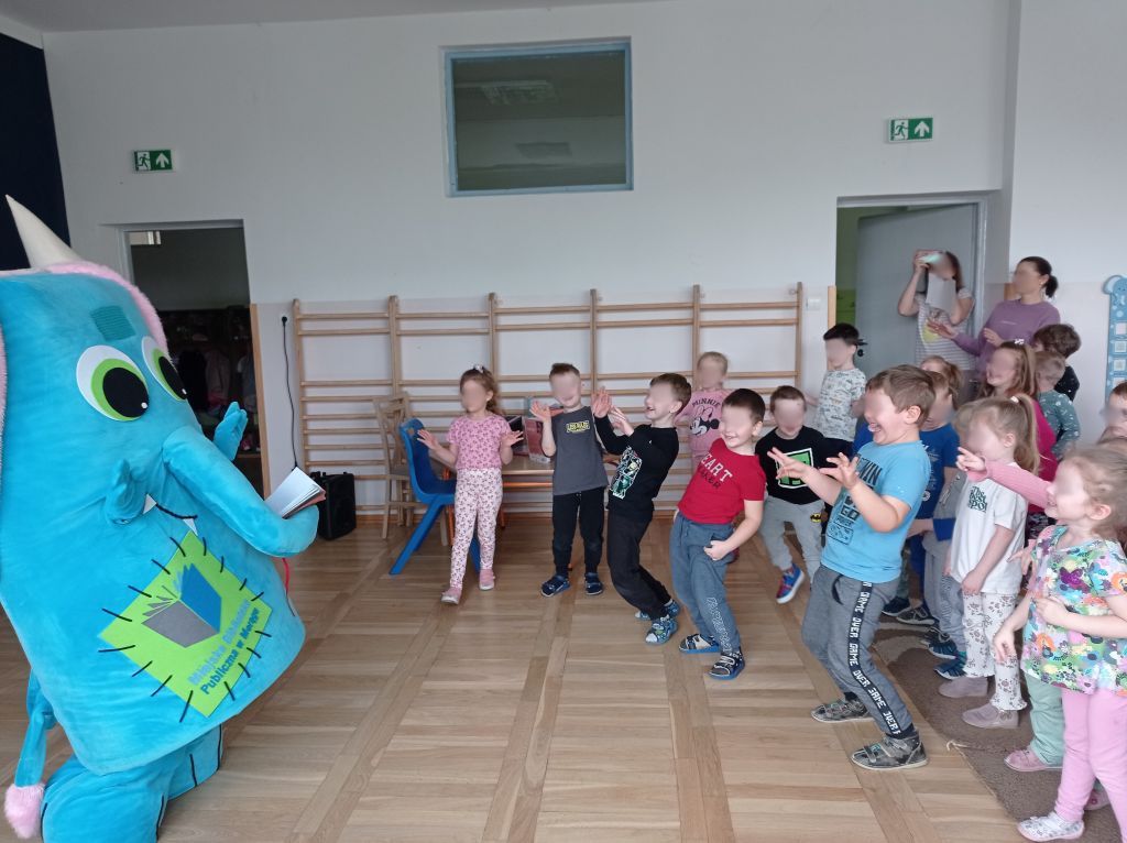 Grupa dzieci tańczy, powtarzając ruchy wielkiej niebieskiej maskotki stojącej naprzeciwko nich