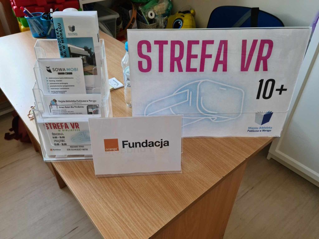 Na biurku stoją ekspozytory, jeden z napisem Strefa VR 10+, drugi z logo fundacji Orange, po lewej stoi ekspozytor z ulotkami