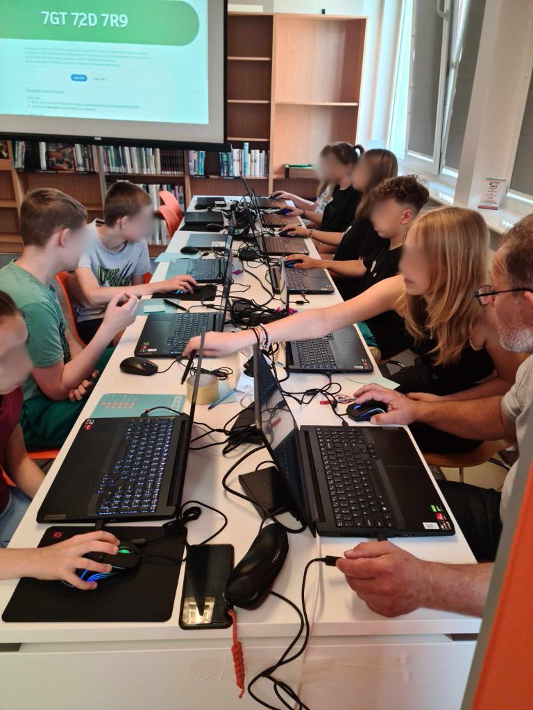 grupa dzieci i nauczyciel siedzą przy laptopach, w tle ekran z wyświetlanym obrazem