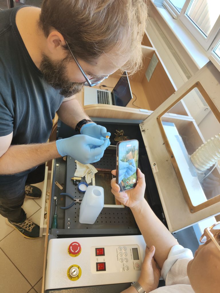 mężczyzna stoi obok plotera laserowa trzymając w rękach mały przedmiot, obok stoi kobieta z wyciągniętą ręką trzymając telefon i nagrywając ręce mężczyzny
