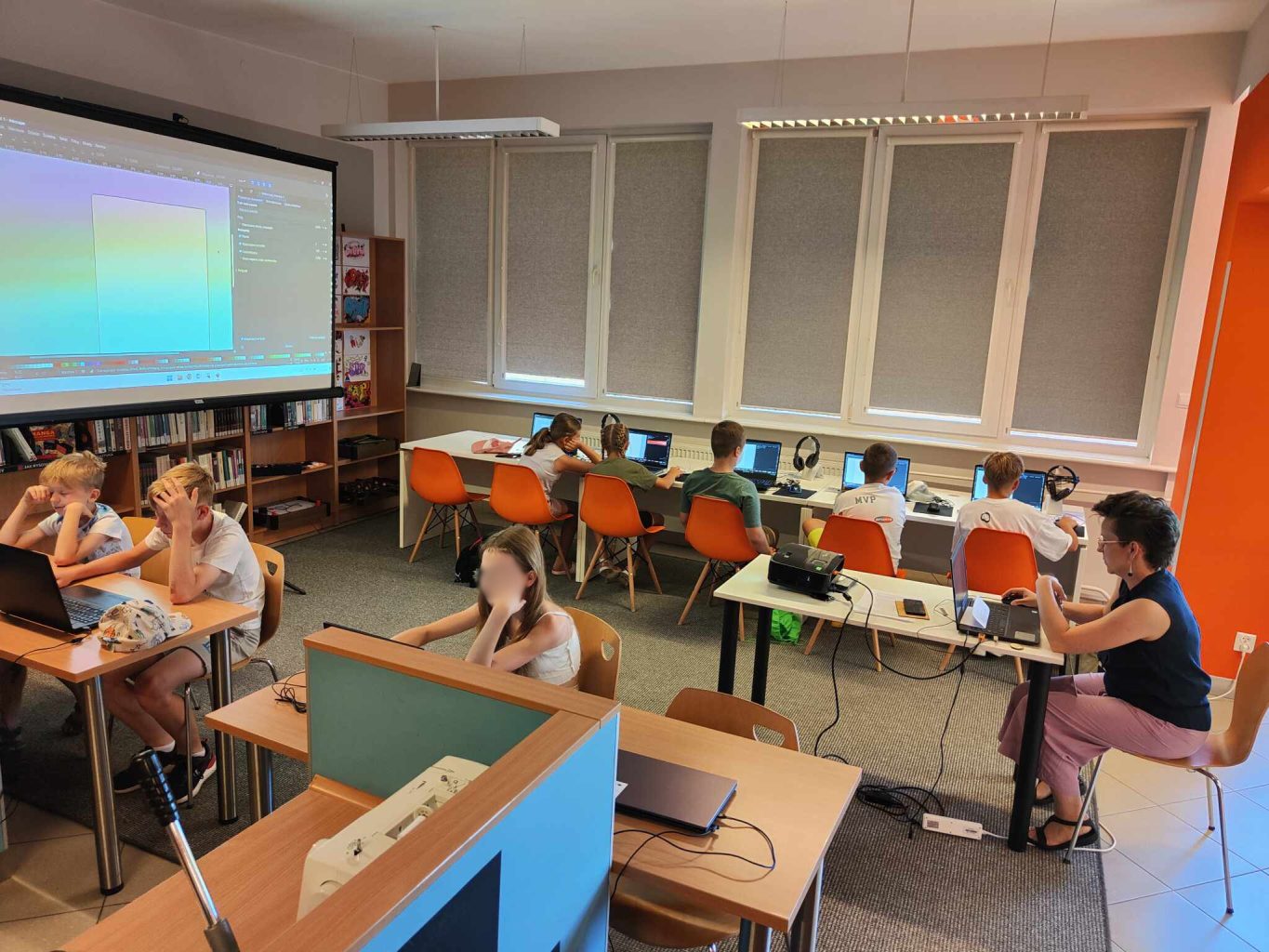 grupa dzieci siedzi przy biurkach przed laptopami, z lewej ekran