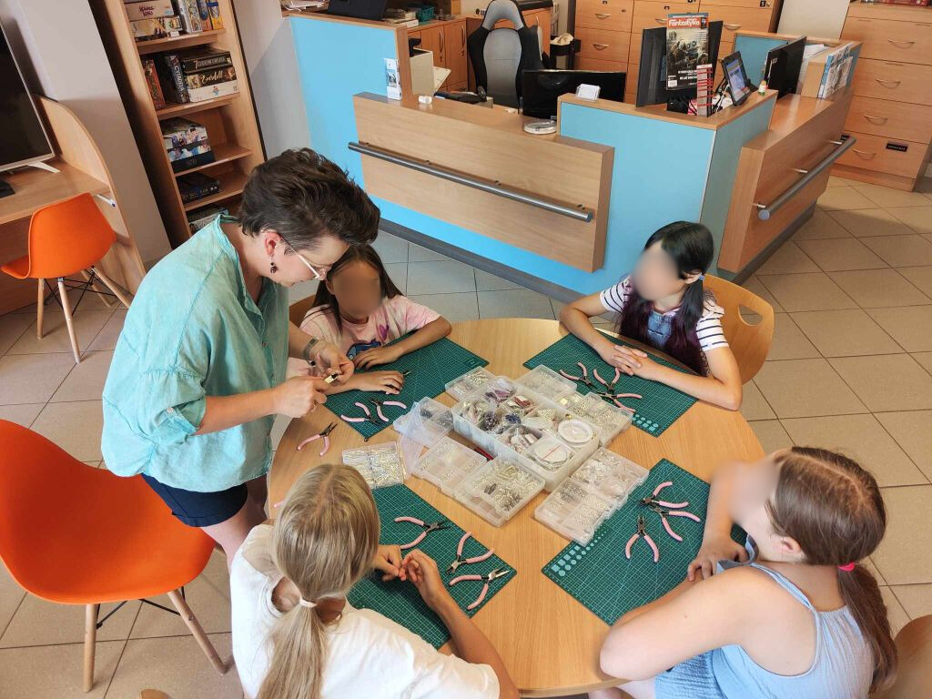 grupa dziewczynek siedzi przy stole na którym leżą maty samogojące oraz narzędzia i pudełka z półproduktami do tworzenia biżuterii, między nimi stoi kobieta prezentująca sposób tworzenia własnych ozdób