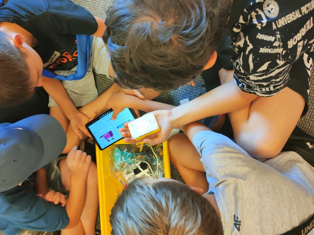 Grupa dzieci siedzi na wykładzinie układając elementy klocków lego. Jeden z chłopców trzyma w rękach tablet i pudełko ze świecącym symbolem.