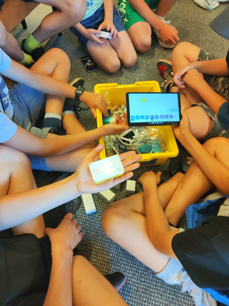 Grupa dzieci siedzi na wykładzinie układając elementy klocków lego. Jedno z dzieci trzyma w ręku tablet a drugie pudełeczko ze świecącym symbolem.