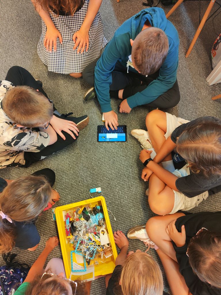 Grupa dzieci siedzi na wykładzinie układając elementy klocków lego. Jeden z chłopców trzyma przed sobą tablet.