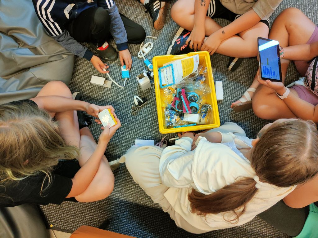 Grupa dzieci siedzi na wykładzinie układając elementy klocków lego. Jedna z dziewczynek trzyma pudełko ze świecącym symbolem a druga tablet.