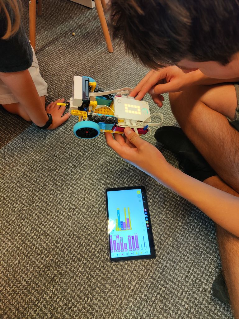 Chłopiec trzyma w ręku robota z klocków lego i pochyla się nad leżącym na podłodze tabletem.
