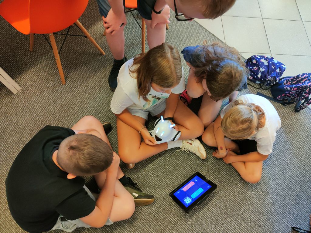 Dzieci siedzą w grupie na wykładzinie, przed nimi stoi tablet i robot Photon