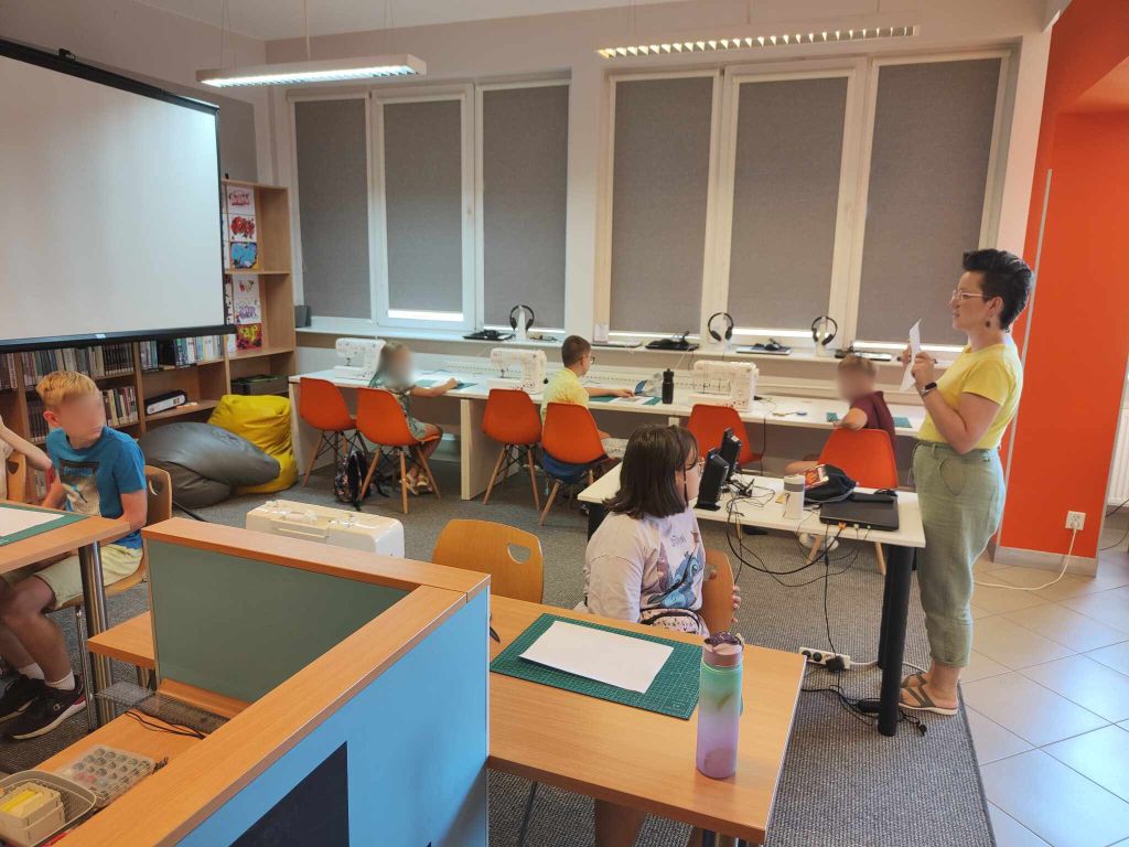 kilkoro dzieci siedzi przy stołach, obok nich stoją maszyny do szycia, po prawej stoi kobieta