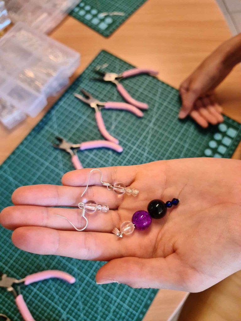 rozłożona dłoń na której leżą własnoręcznie zrobione kolczyki w różnych kolorach i kształtach, w tle maty samogojące oraz narzędzia i pudełka z półproduktami do tworzenia biżuterii